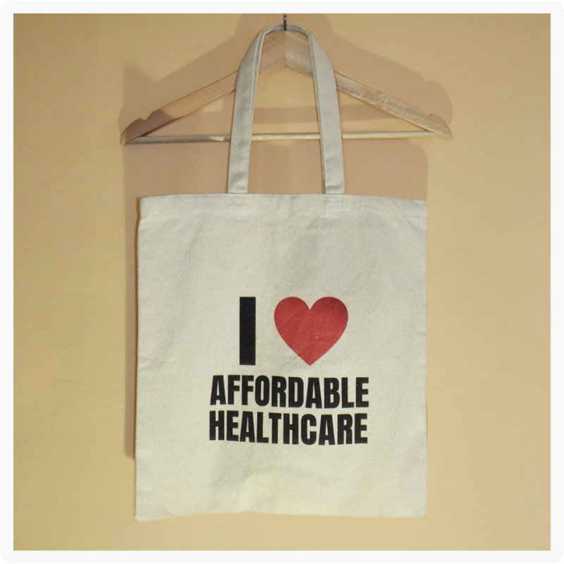 I Love Affordable Healthcare canvas tote bag with red heart and bold black lettering hanging on a wooden hanger